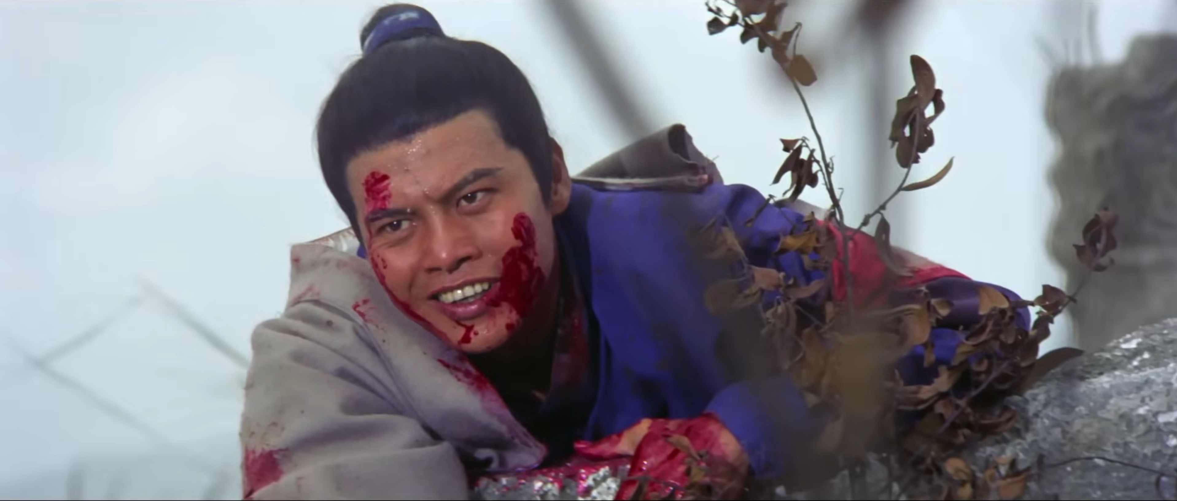 Lo Lieh, dressed in a blue robe and covered in blood, leans over a rock. He's smiling at something unseen.