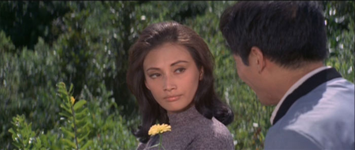 Jenny Hu, dressed in a grey turtleneck, stands in a field and looks at a man with his back to the camera. She looks disappointed or angry.