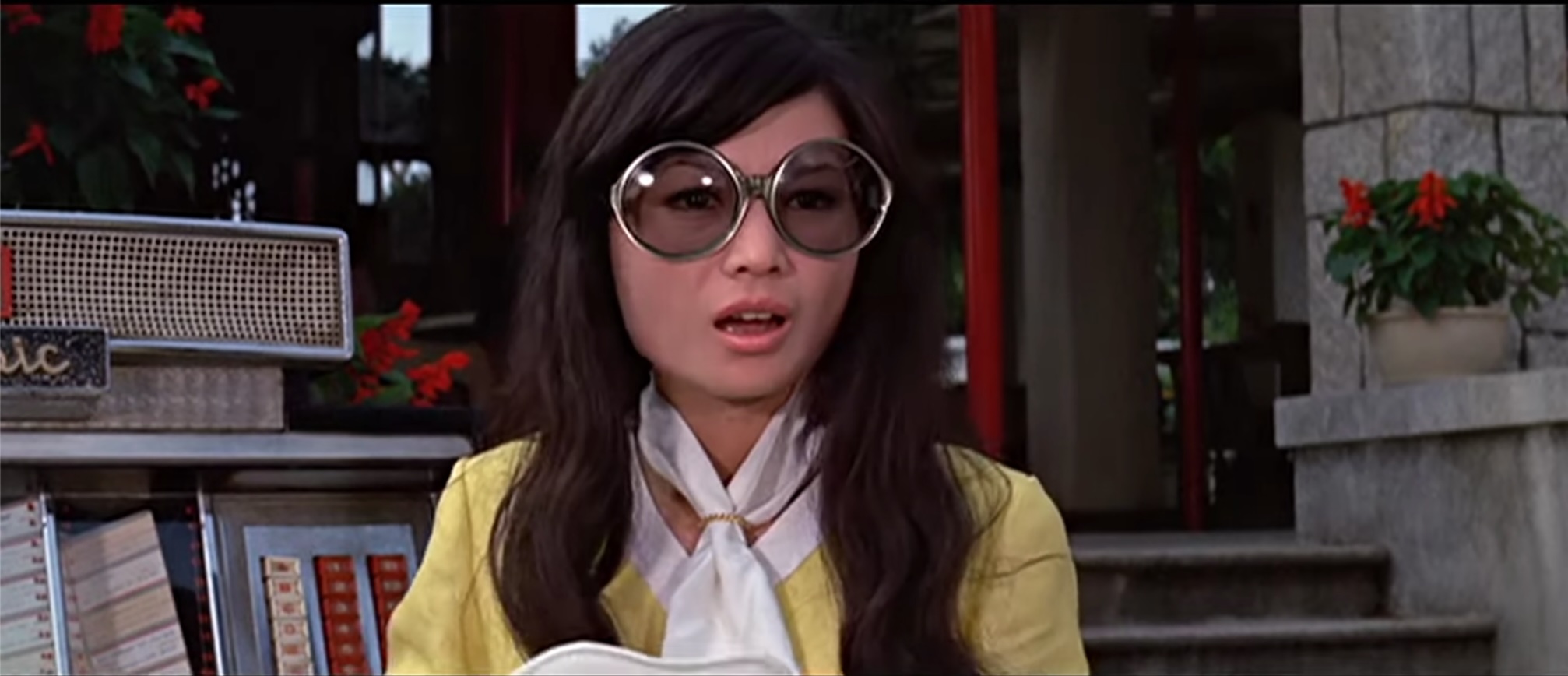 A close up of Ching Li, wearing a yellow and white mini dress and massive gold-rimmed sunglasses. She's looking surprised.