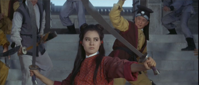 A young woman wearing a red and gold top stands in front of several armed men. She is wielding two swords. The man directly behind her has his arm raised as if to strike.