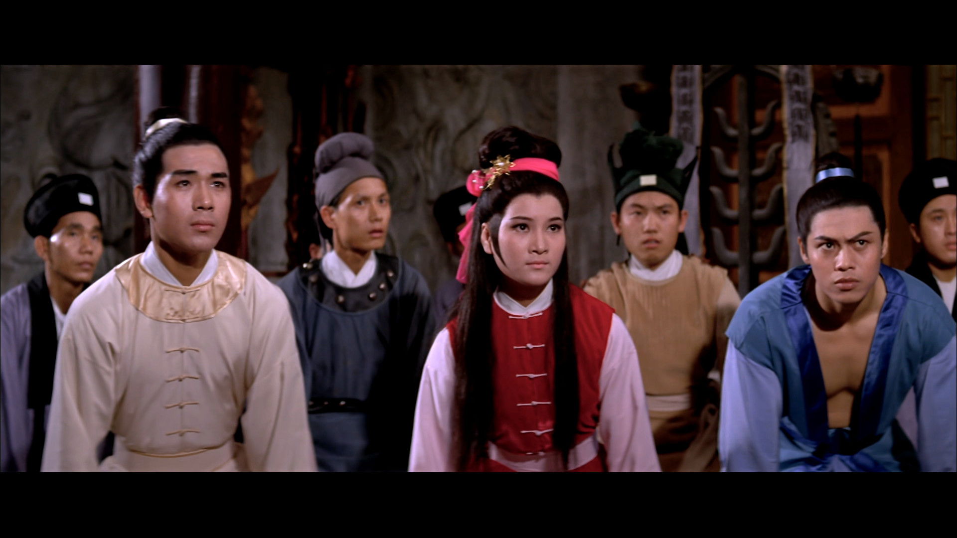 Shu Pei-Pei wearing a red and white tunic kneels on the floor. Around her, also kneeling, are six men.